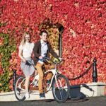 Young_happy_tourist_couple,_handsome_bearded_man_and_pretty_long-haired_blond_woman_cycling_tandem_bicycle_on_bright_sunny_day_on_background_of_building_fully_overgrown_with_red_ivy_leaves.