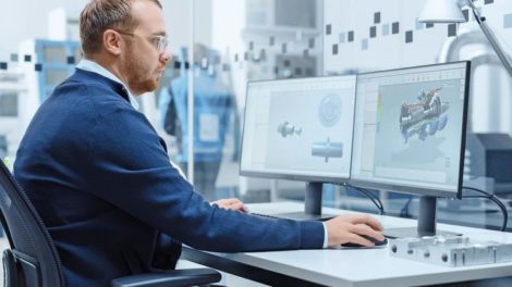 Male_Industrial_Engineer_Solving_Problems,_Working_on_a_Personal_Computer,_Two_Monitor_Screens_Show_CAD_Software_with_3D_Prototype_of_Hybrid_Electric_Engine_Being_Tested._Working_Modern_Factory