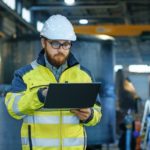 Industrial_Engineer_in_Hard_Hat_Wearing_Safety_Jacket_Uses_Laptop._He_Works_in_the_Heavy_Industry_Manufacturing_Factory_with_Various_Metalworking_Processes_are_in_Progress.