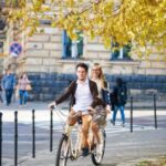 Young_handsome_man_and_pretty_blond_woman_riding_tandem_double_bicycle_along_paved_sidewalk_under_golden_yellow_trees_on_bright_sunny_autumn_day_on_stone_old_building_and_walking_people_background.